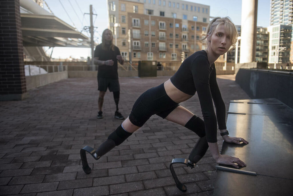 Blonde woman standing outside wearing her running blades stretching and looking straight at camera