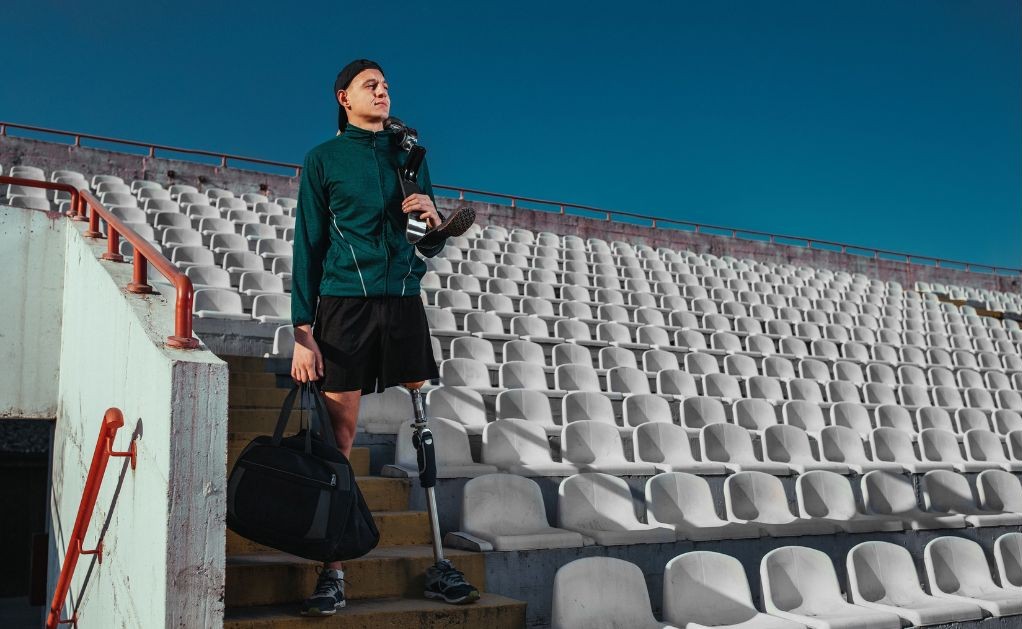 A young athlete standing on stadium steps with a prosthetic leg, holding a gym bag and a pair of shoes.