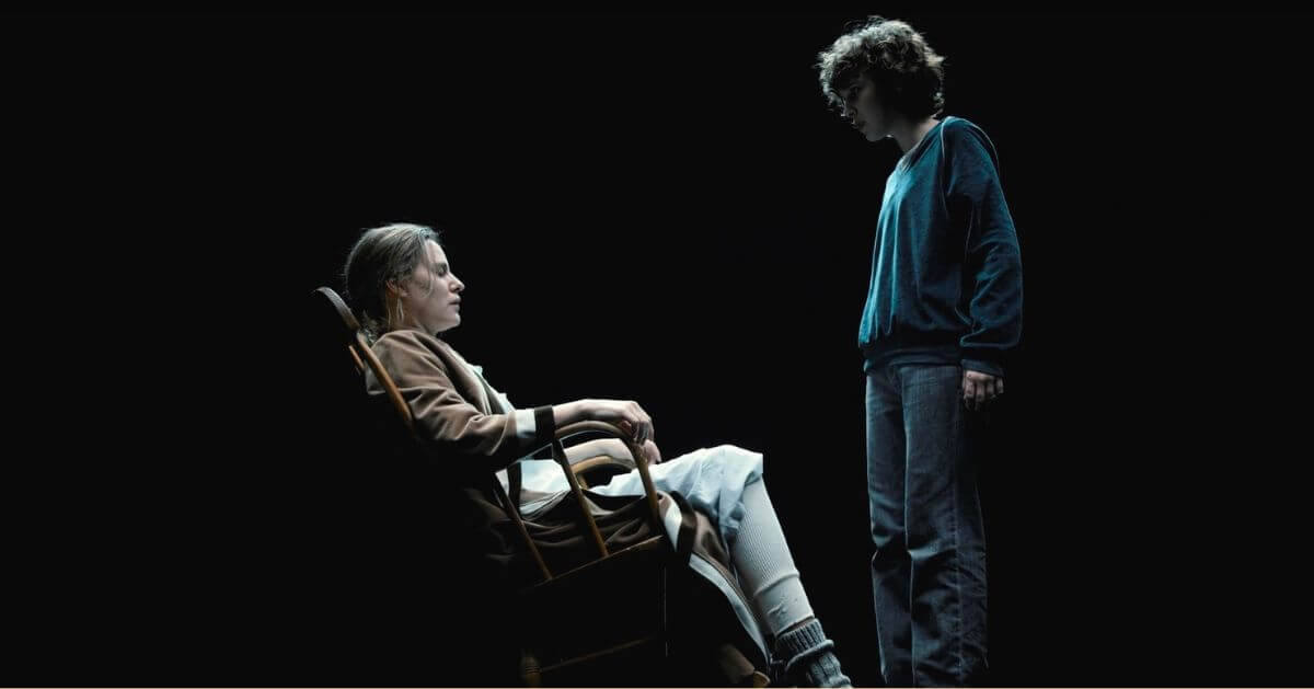 A scene from Stranger Things series featuring Aimee Mullins sitting in a rocking chair facing Millie Bobby Brown, who stands in front of her, looking down.