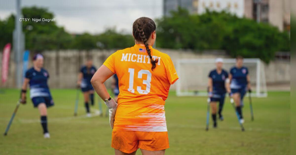 Alexia Michitti, wearing an orange jersey with the number 13, stands on a soccer field as a goalkeeper.