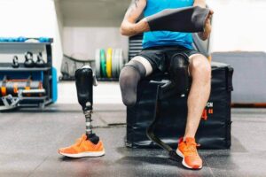 A man with a leg amputation sits on a gym box while putting on a prosthetic socket liner. His running blade and prosthetic leg with a sneaker are placed in front of him on the gym floor.