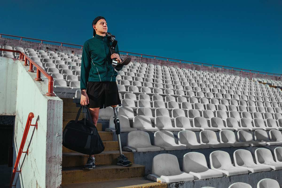 An athlete with a prosthetic leg walks down stadium stairs carrying a gym bag and a running blade.