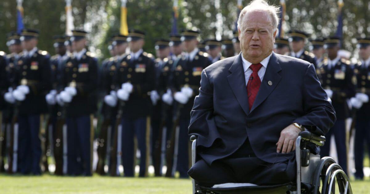 Max Cleland, a U.S. Senator and war veteran, is shown sitting in a wheelchair during a formal ceremony.
