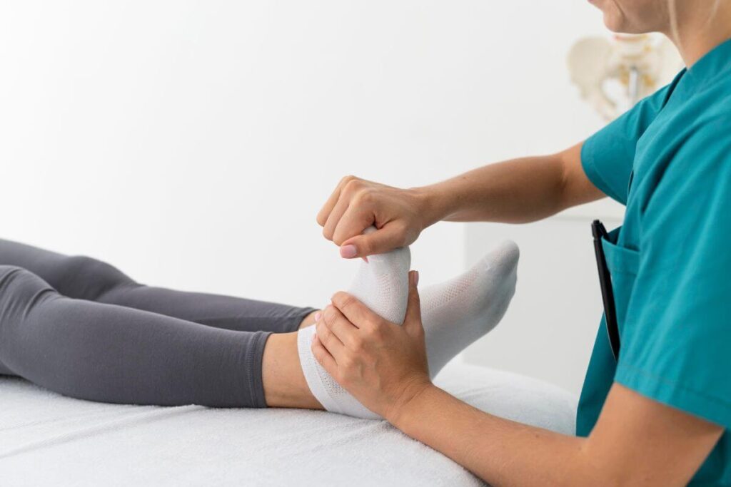 A healthcare professional in teal scrubs stretches the foot of a person lying down during a physical therapy session.