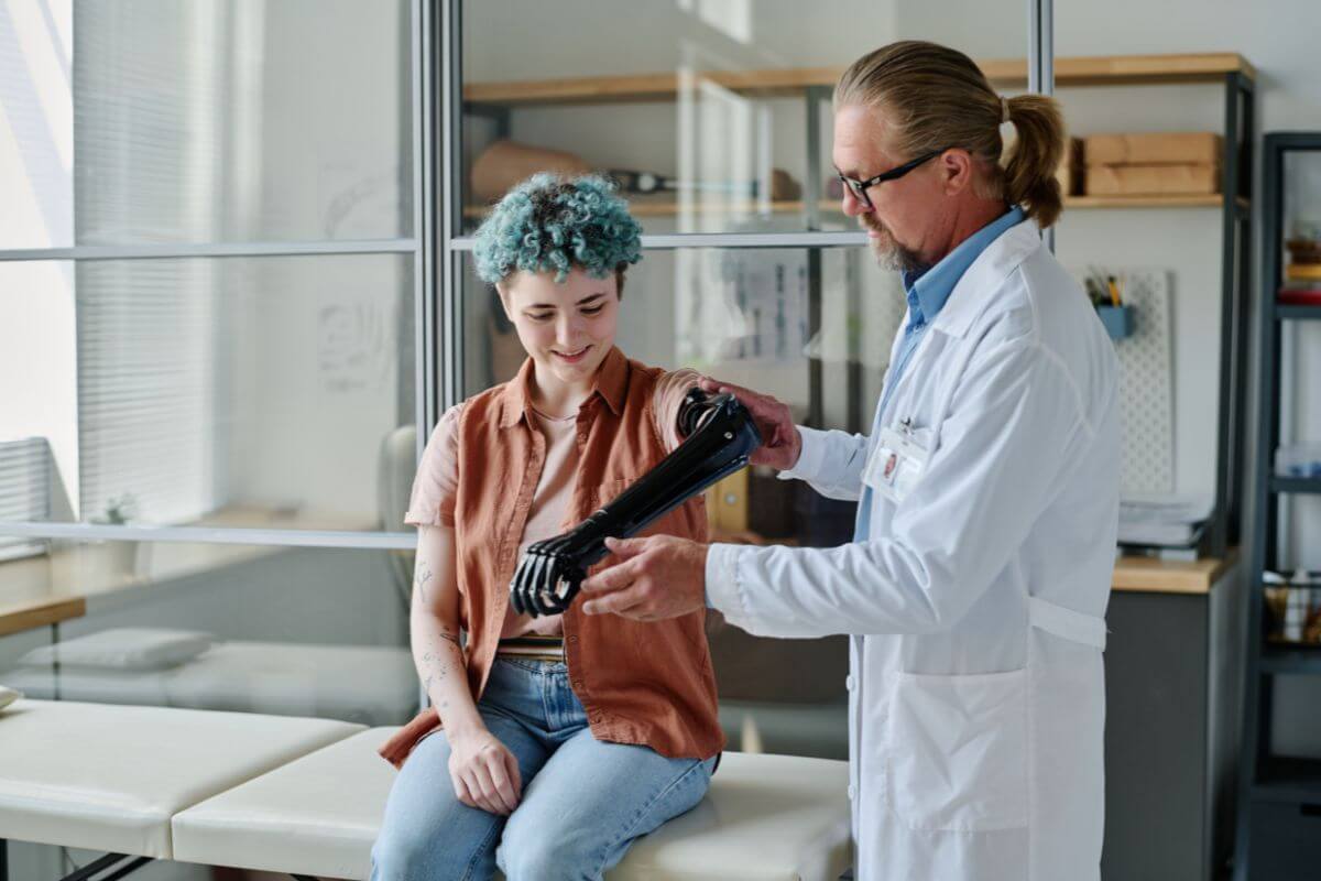 A medical professional helps a young woman try on a black prosthetic arm in a clinical setting.