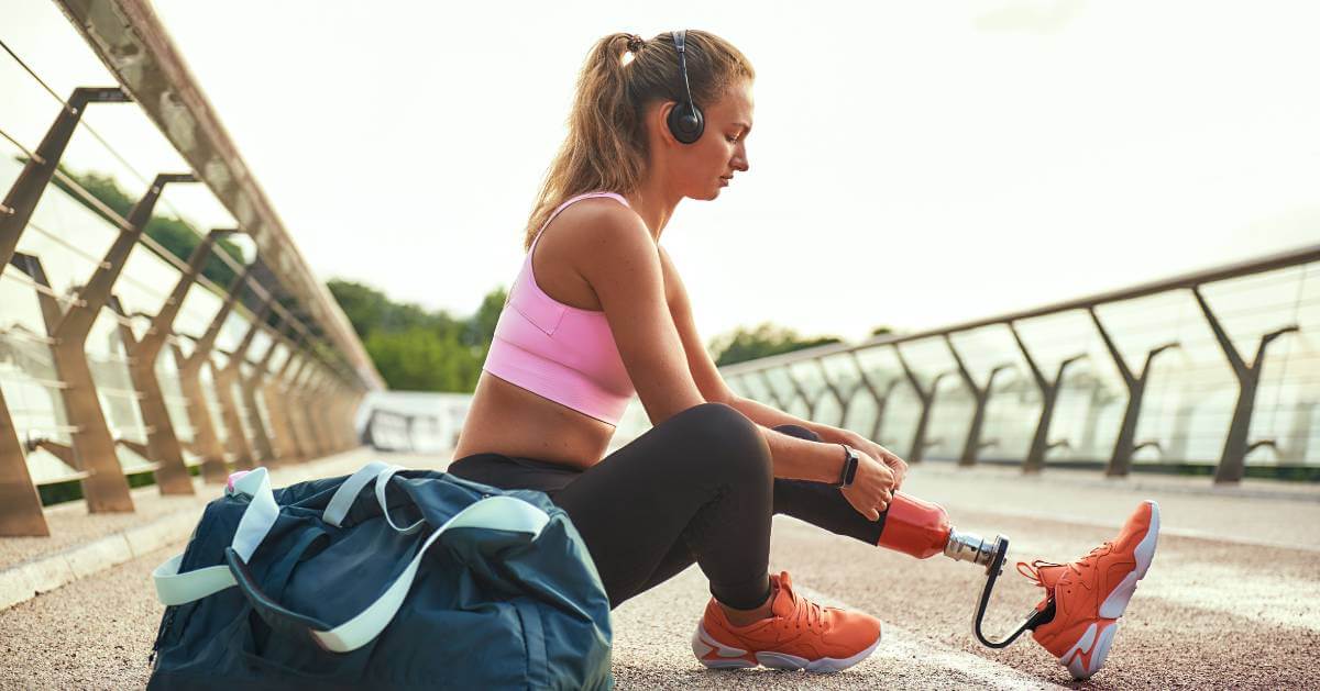 A woman wearing sports attire and headphones, adjusting her prosthetic leg on the ground