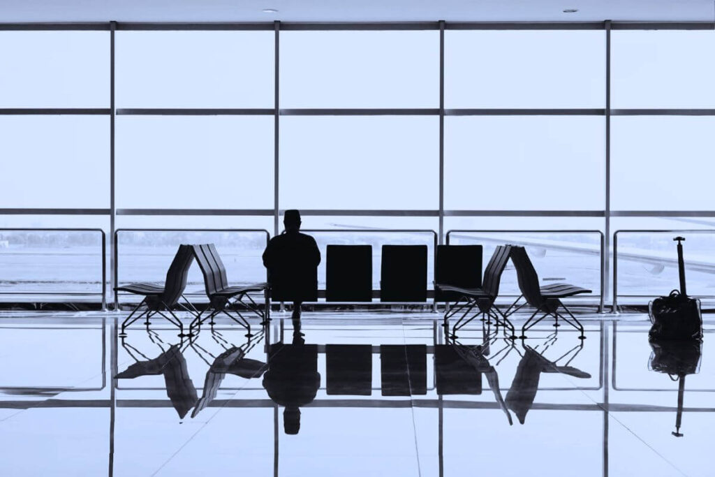Silhouette of a person sitting alone in an empty airport terminal, with rows of seats and a travel bag near a large window overlooking the runway.