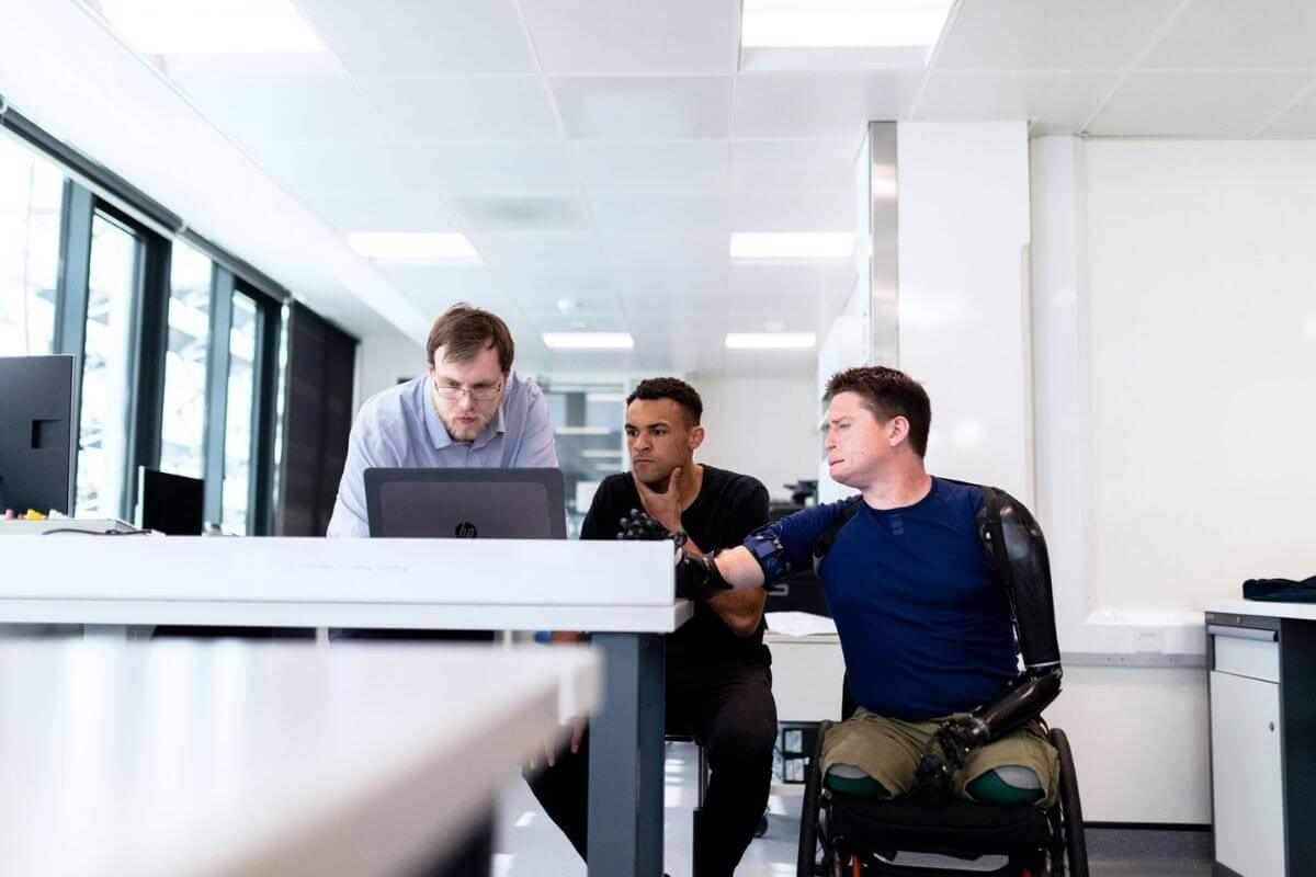Man with bilateral prosthetic arms in a wheelchair with two men around a laptop in an office.