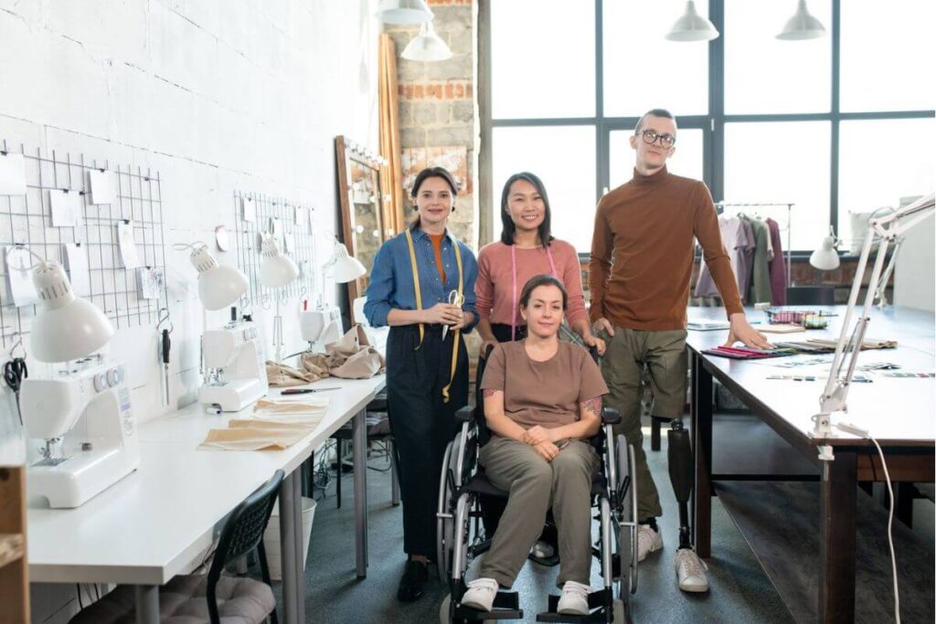 Inclusive workplace with a small fashion design team standing beside a colleague using a wheelchair and prosthetic leg inside a bright sewing studio.