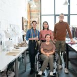 Inclusive workplace with a small fashion design team standing beside a colleague using a wheelchair and prosthetic leg inside a bright sewing studio.