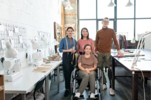 Inclusive workplace with a small fashion design team standing beside a colleague using a wheelchair and prosthetic leg inside a bright sewing studio.