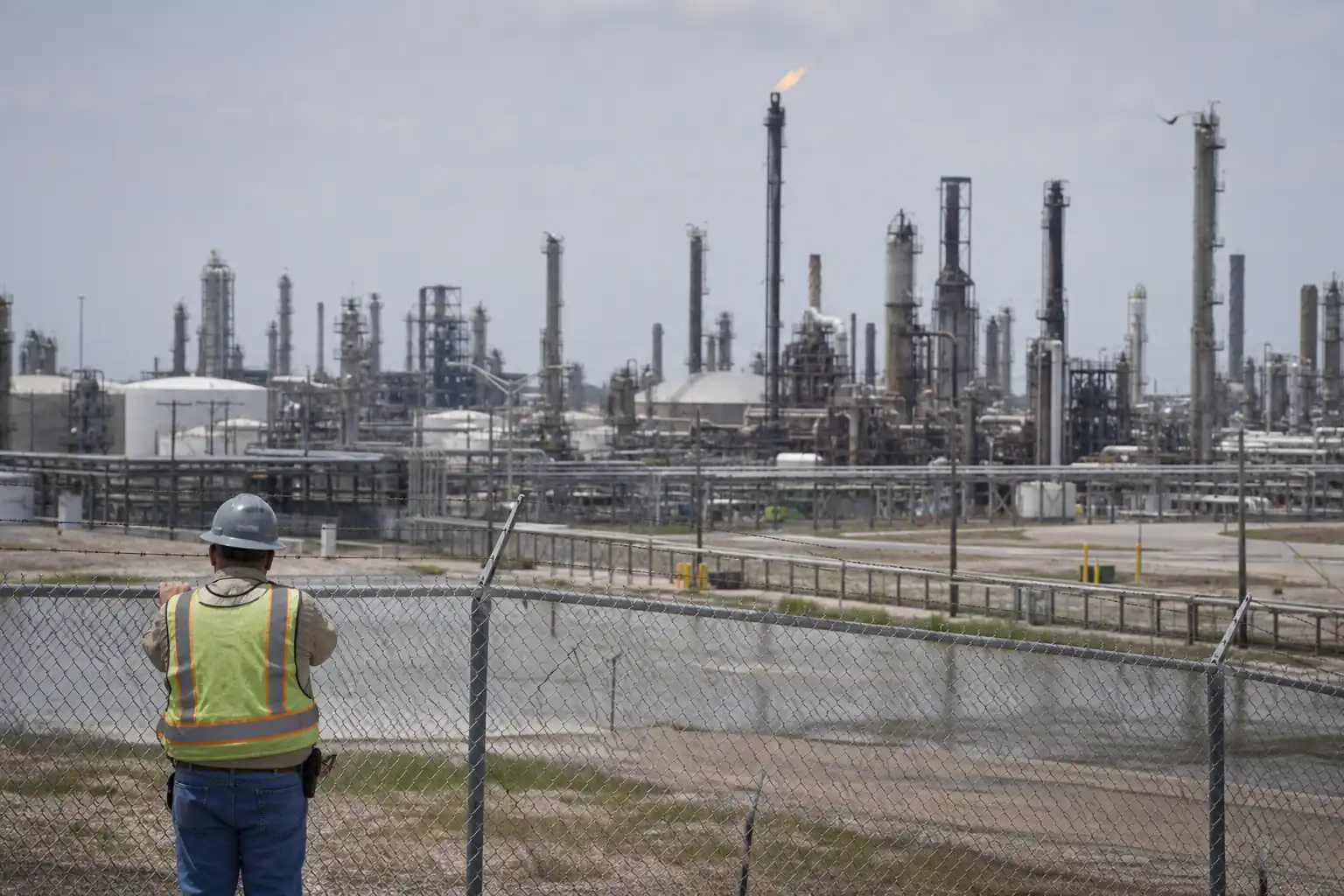 Vast Houston Ship Channel petrochemical refinery with distillation towers, storage tanks, piping infrastructure, and a flare stack visible behind a chain-link fence with a worker in hard hat and hi-vis vest in the foreground