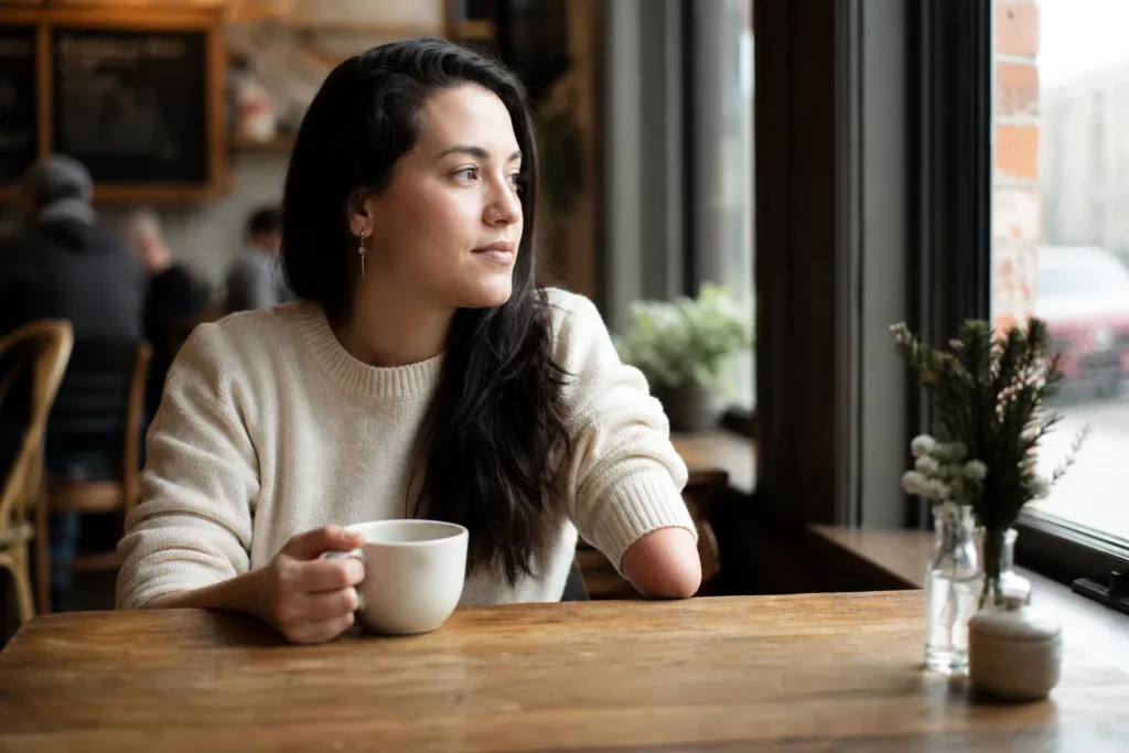 Young adult woman with an upper-limb difference sitting thoughtfully at a cafe table holding a coffee mug