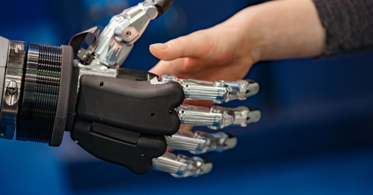 Close-up of a mechanical prosthetic hand reaching toward a person’s hand.