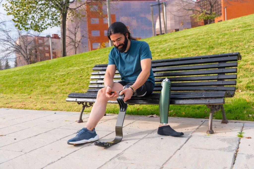 Person adjusting a below-knee prosthetic leg while sitting on a park bench outdoors.