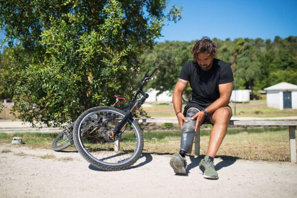 Man with a prosthetic leg adjusting his prosthesis while sitting beside a mountain bike outdoors.