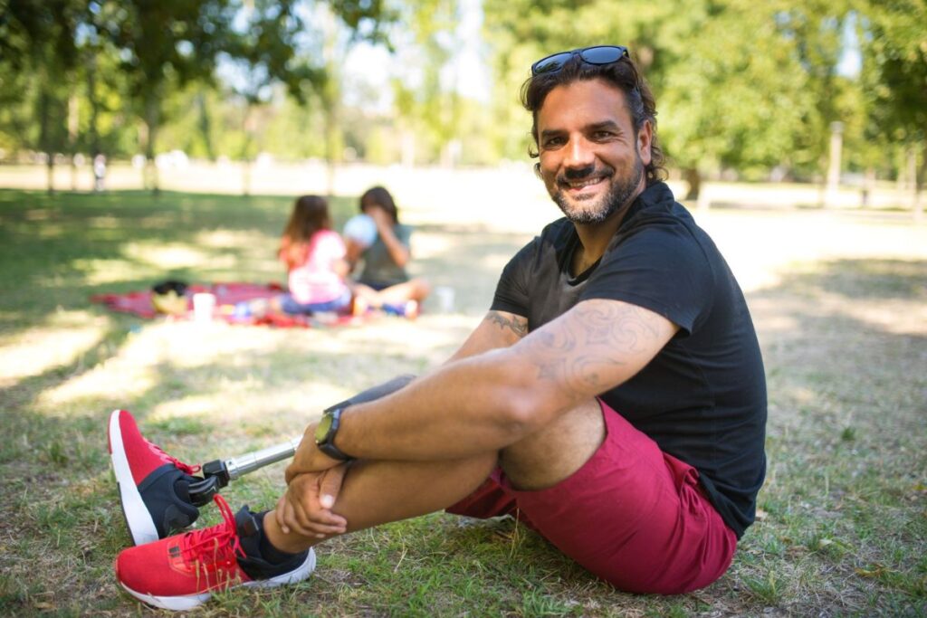 Person with a prosthetic leg sitting in a park with family in the background on a sunny day.