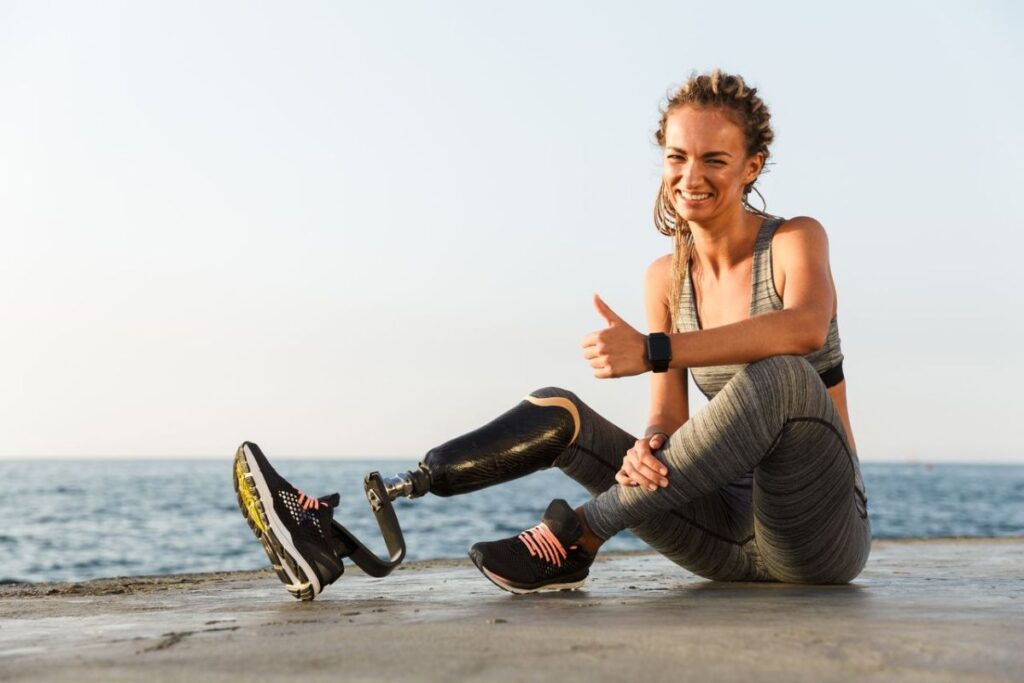 Woman with a below-knee running prosthesis sitting by the waterfront and giving a thumbs-up after a workout.