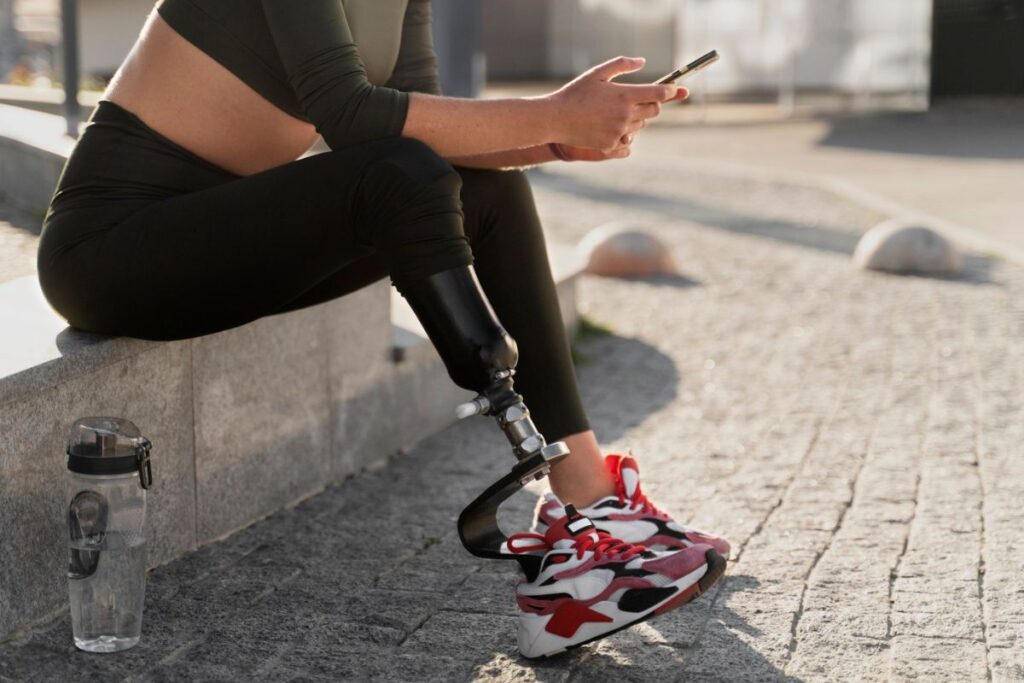Person with a below-knee prosthetic leg sitting outdoors in athletic clothing and sneakers while using a smartphone.