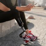 Person with a below-knee prosthetic leg sitting outdoors in athletic clothing and sneakers while using a smartphone.