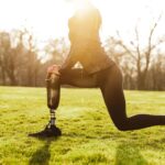 Woman with a prosthetic leg stretching outdoors on grass.